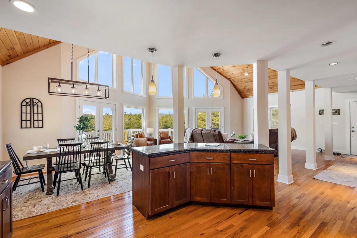 A cheerful, open dining area anchored by a large table, with a view into the kitchen. Sunlight pours in through expansive windows, illuminating the wooden beams and modern kitchen island, creating a vibrant atmosphere ideal for shared meals and festivities.