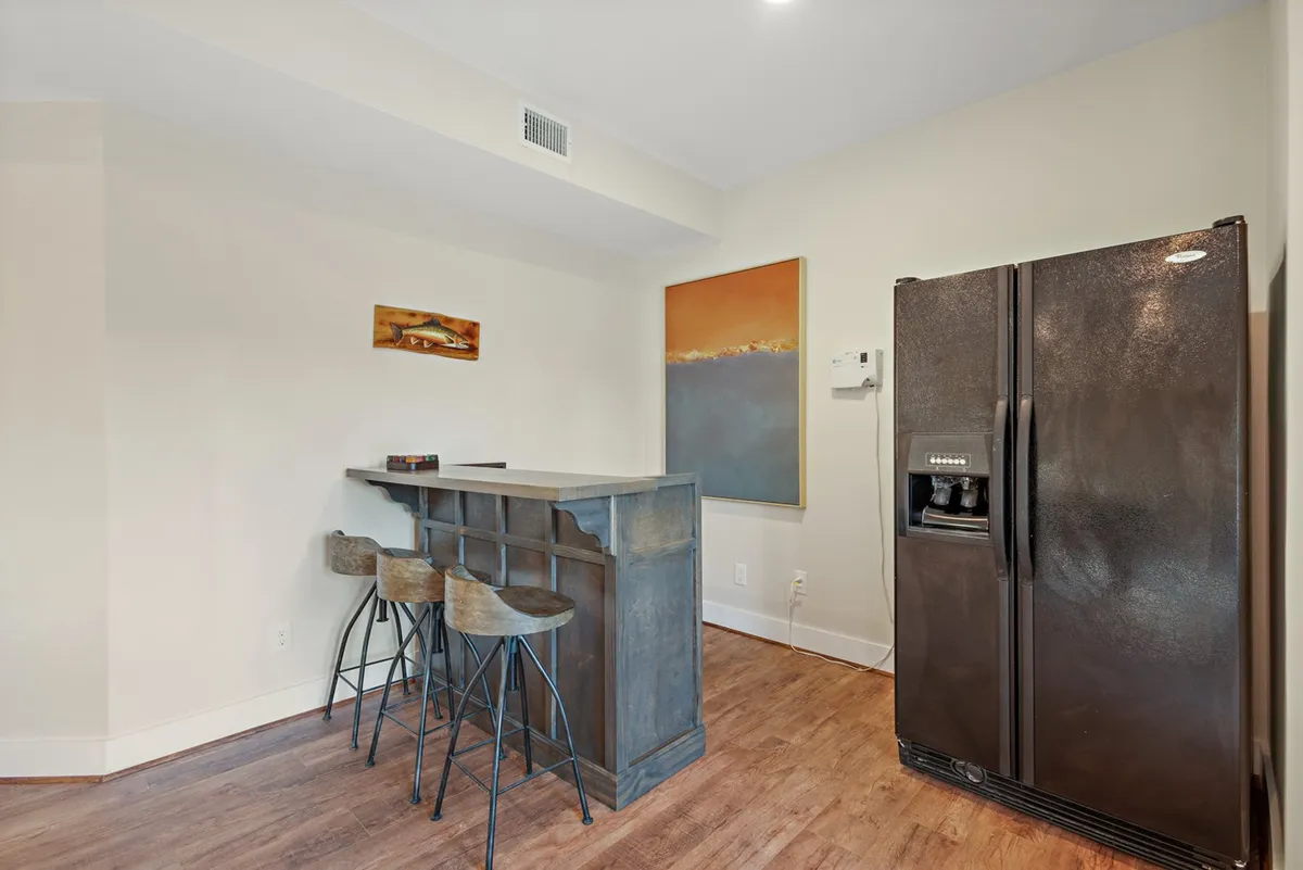 A stylish wet bar and bar counter area featuring a sleek refrigerator, decorative artwork, and comfortable seating. The scene suggests a place for casual dining, mixing drinks, and socializing after a round of games or before a movie.