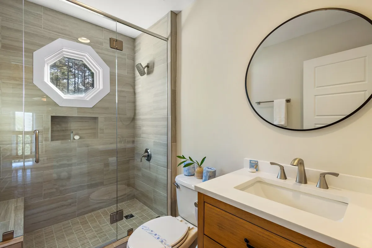 A modern bathroom with a glass shower enclosure, an octagonal window that lets in natural light, a round mirror, and a sleek vanity sink. The space feels bright, clean, and functional.