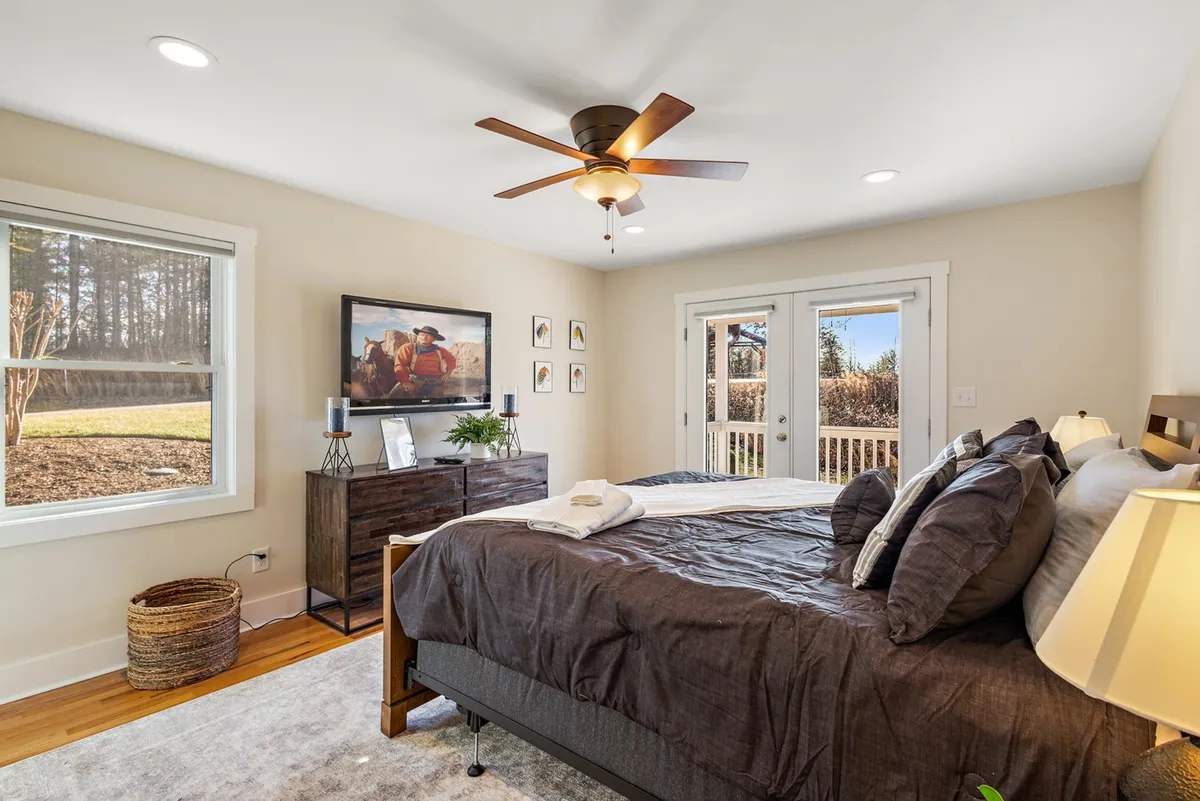 A tranquil bedroom featuring a king-sized bed, a dresser, TV, sliding doors leading to a private deck, and tasteful wall art. Natural light fills the space, creating a soothing and inviting atmosphere—ideal for relaxation or quiet mornings.