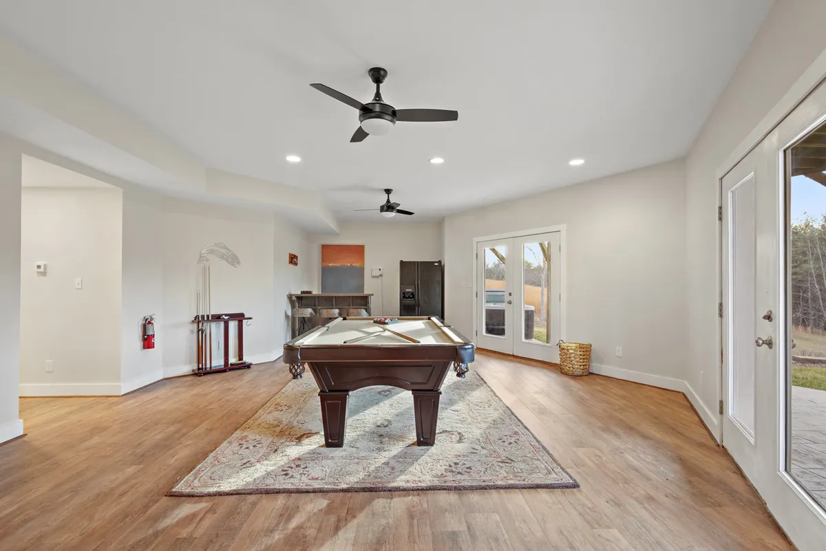 A lively lower-level recreation space featuring a pool table atop a patterned rug, with ample natural light streaming through large windows. The scene invites playful gatherings, relaxation, and friendly competitions, embodying the home's entertainment-focused amenities.
