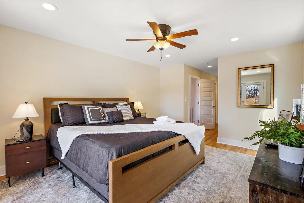 A tranquil queen bedroom with a neatly made bed, nightstands, a mirror, and lush plants, all bathed in natural light. The layout is simple yet inviting, perfect for restful nights after a day of group activities.