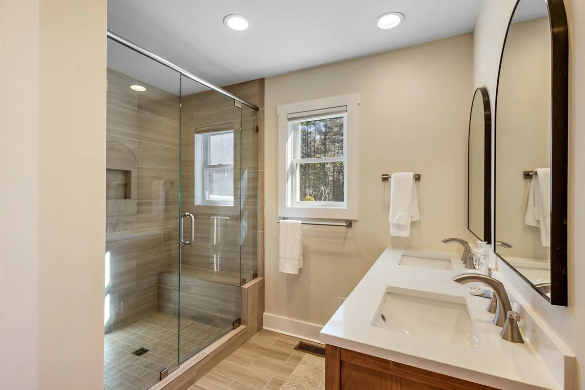 A modern, light-filled bathroom with a glass shower enclosure, double vanity, a large mirror, and a window letting in natural light. The inviting ambiance and ample space suit both leisurely routines and busy mornings.