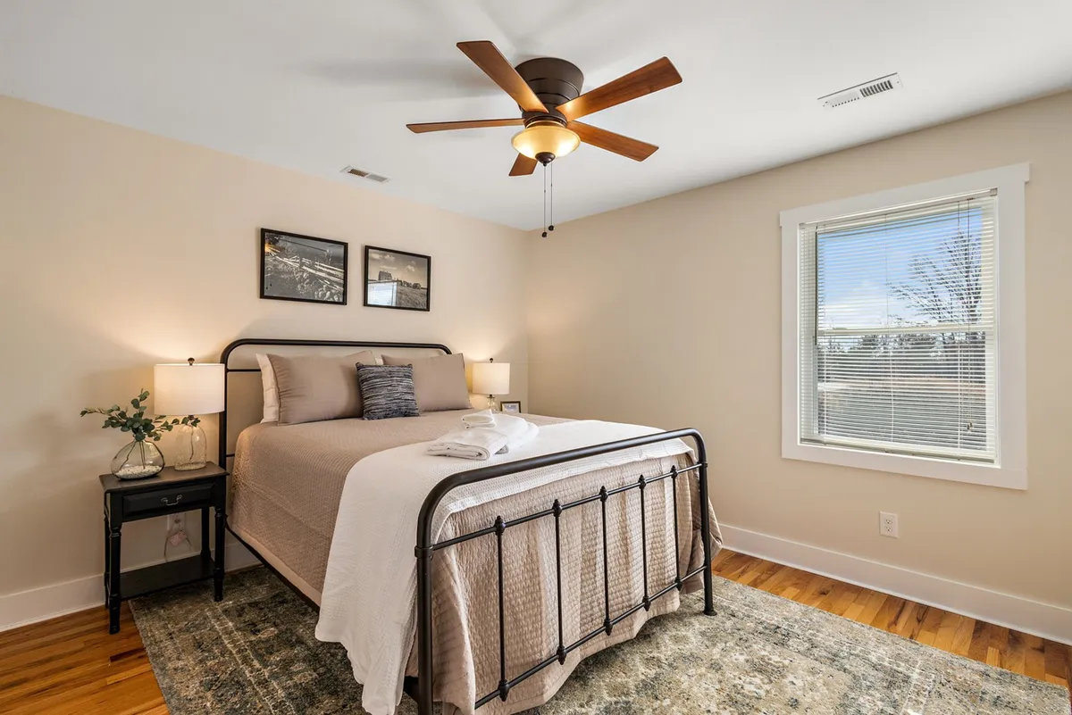 A tranquil queen bedroom with a plush bed, nightstand, ceiling fan, window, and framed pictures. Soft natural light streams in, creating a peaceful and private atmosphere ideal for restful sleep or quiet moments alone.