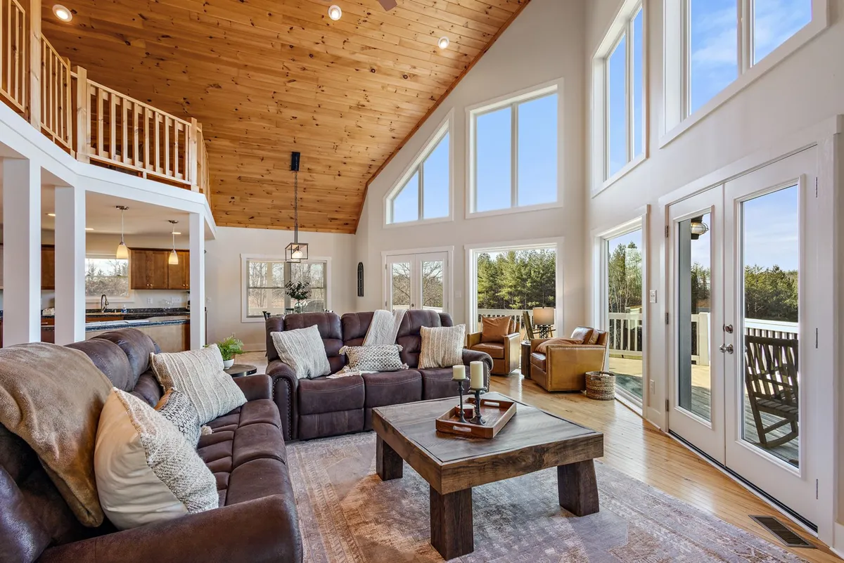 A welcoming loft lounge space with large windows that flood the room with natural light. The area features comfortable seating and a coffee table, all set beneath a warm wooden ceiling. The arrangement invites relaxation, reading, and quiet conversation, and the expansive windows offer inviting views.